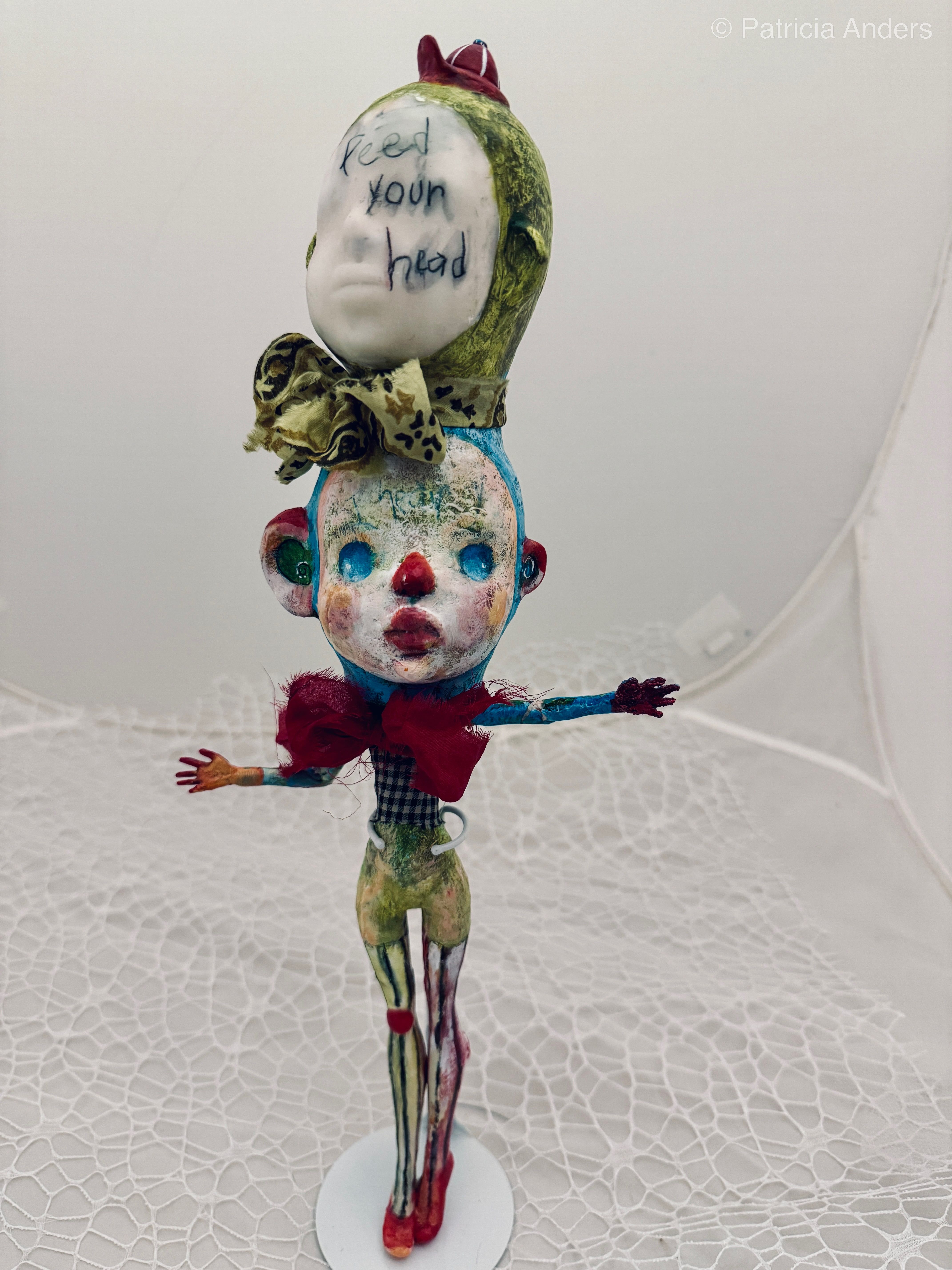 A mixed media sculpture featuring two stacked doll-like heads with a figure body below, decorated with fabric, paint, and text reading "feed your head," standing approximately 16 inches tall with outstretched mismatched arms.