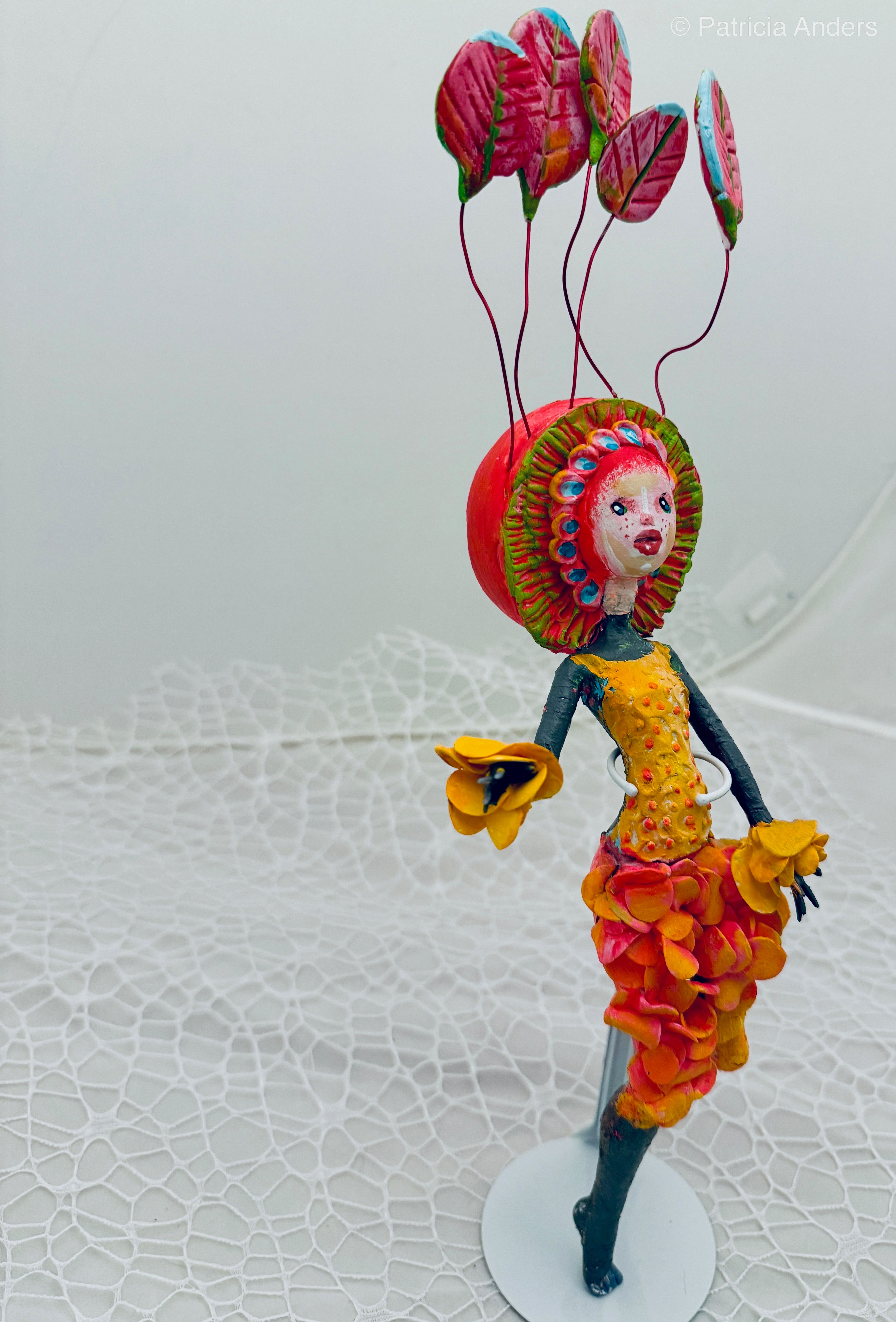 A whimsical mixed media sculpture of a figure with a red mushroom cap crowned by pink leaves on wire stems, wearing a dress made of layered yellow and pink petals with blue-gray limbs.
